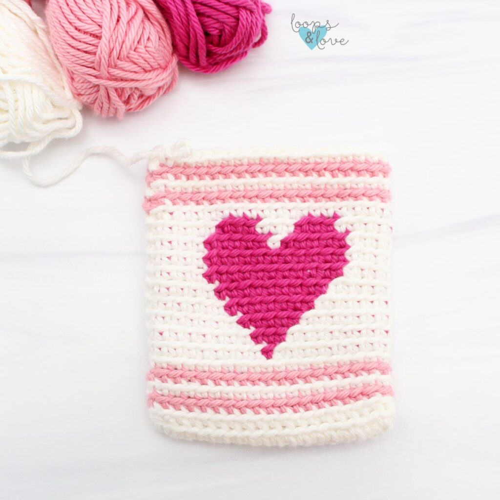 White crochet drawstring bag with a pink heart, with balls of white and pink yarn.