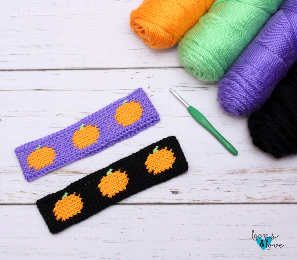 Two crochet pumpkin ear warmers in purple and black with orange pumpkin motifs, with yarn and a crochet hook.
